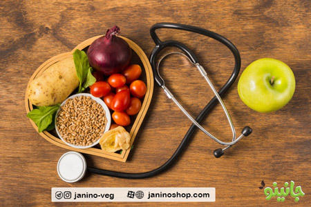 گوشی پزشکی و ظرفی به شکل قلب حاوی سبزیجات، حبوبات، دانه ها، میوه و یک سیب سبز در کنار گوشی پزشکی
