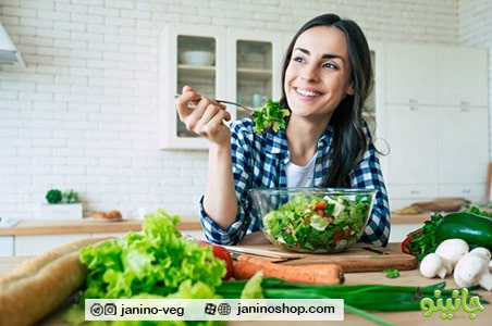 خانمی در محیط آشپزخانه ای با دیوار و کابینت های سفید و پشت کانتر در حال خوردن سالاد سبزیجات روی کانتر سبزیجات متنوع، قارچ، تخته برش آشپزخانه وجود دارد
